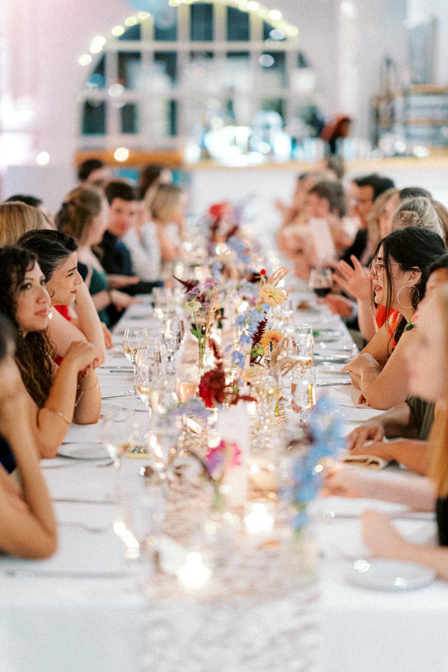 Elegant dinner party with guests seated at a long table, adorned with colorful flowers and glowing candles in a softly lit room.