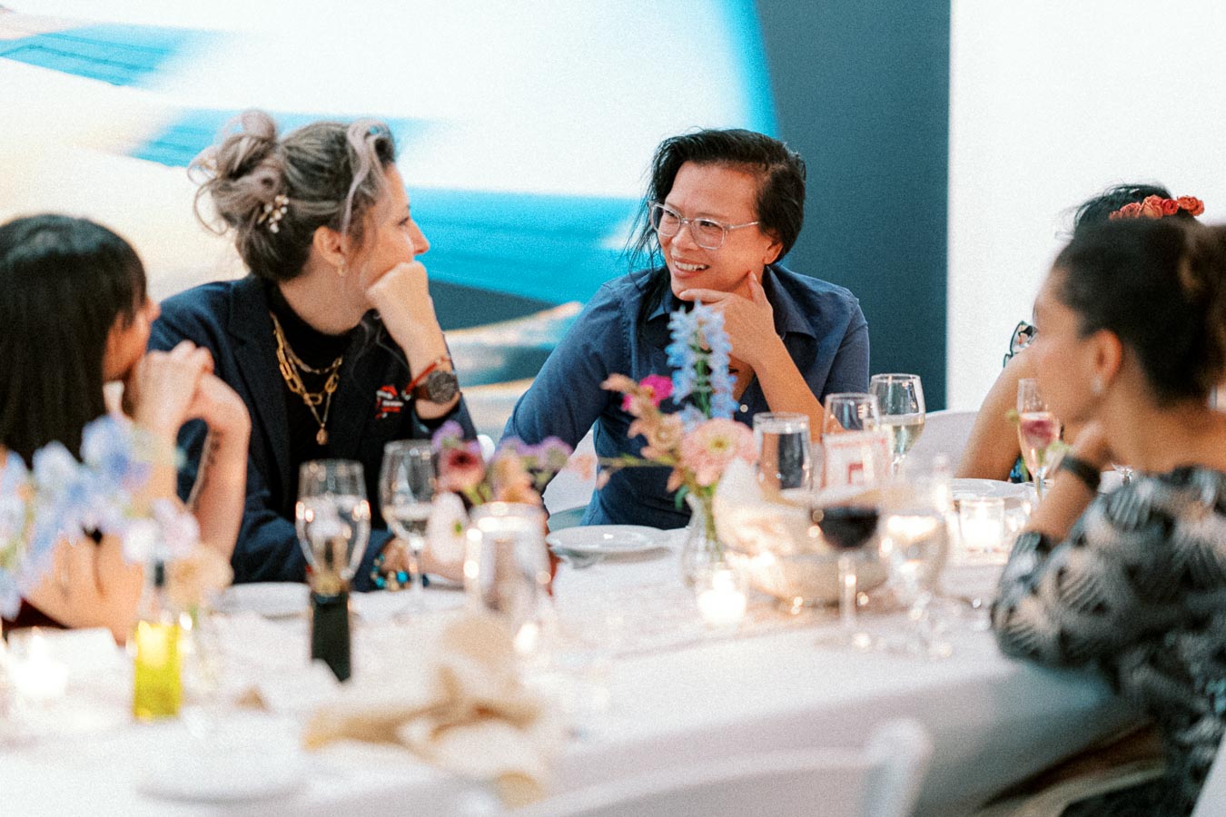 Group of people smiling and conversing at an elegantly set dinner table with flowers and candles, creating a warm and inviting atmosphere.