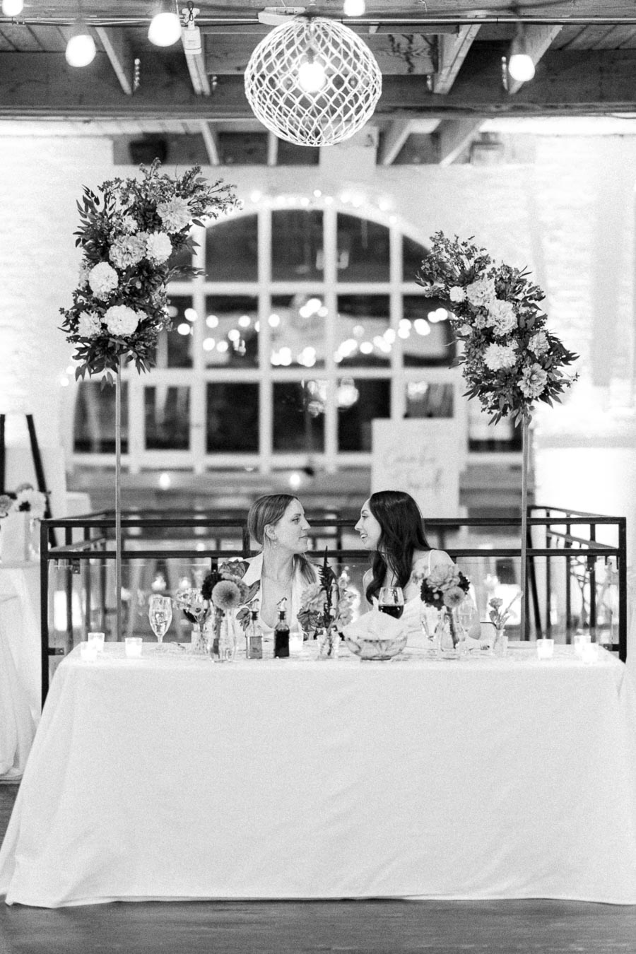 A black and white image of two women seated at a beautifully decorated table adorned with floral arrangements, elegant tableware, and dim lighting, creating a romantic atmosphere. They are looking at each other under a stylish hanging lamp, with a backdrop of large windows and soft, glowing lights.