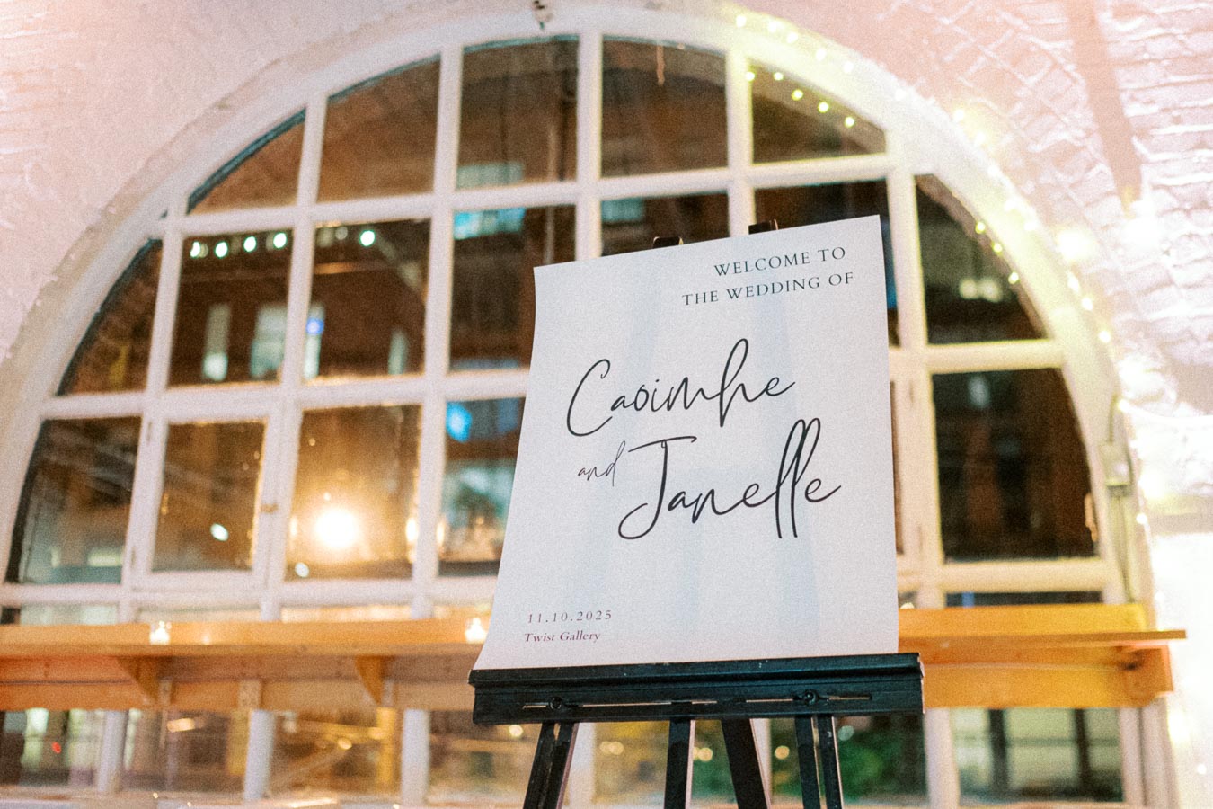 Wedding welcome sign on an easel, featuring elegant calligraphy for Caoimhe and Janelle's ceremony at Twist Gallery on November 10, 2025, with a softly lit, arched window in the background.