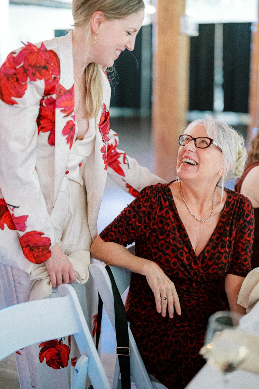 A woman in a floral-patterned suit smiling and interacting with an elderly woman in a patterned dress at a social event.