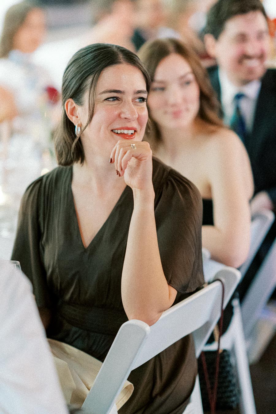 A person sitting at an event, smiling and appearing engaged, with other attendees visible in the background.