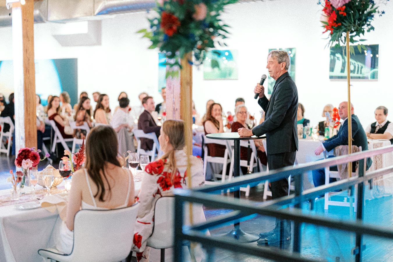 A man in a suit speaks into a microphone at an elegantly decorated wedding reception, with guests seated at round tables adorned with flowers and candles.