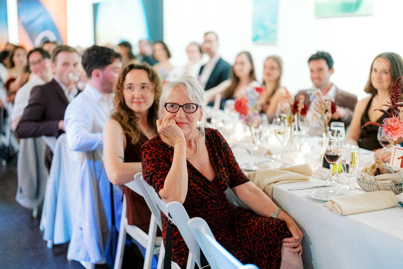 A group of elegantly dressed guests seated at a long table during a formal event, featuring floral arrangements and table settings, creating a sophisticated atmosphere.