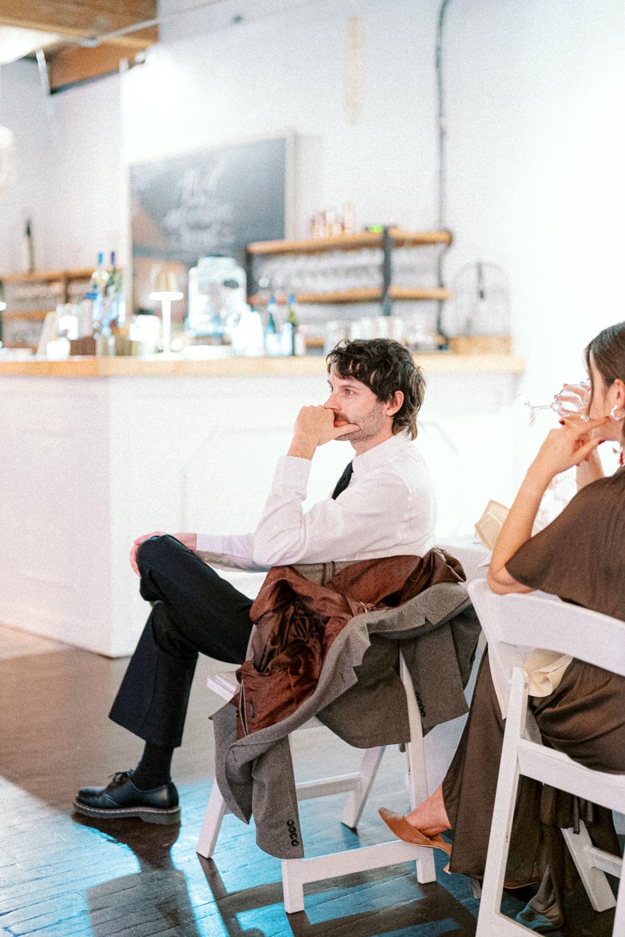 Man in formal attire attentively seated at a stylish, modern event venue with a rustic bar background.