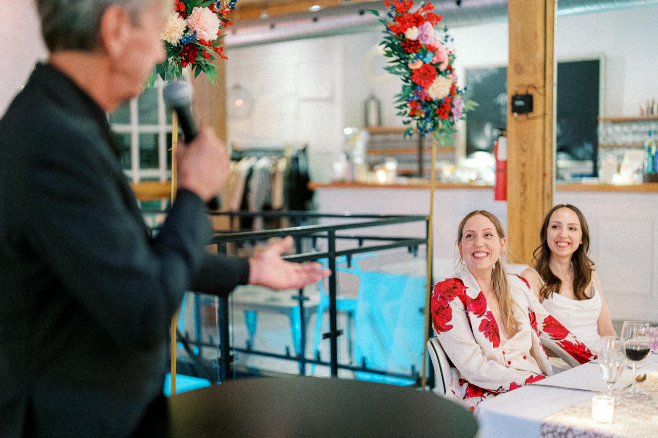 Wedding speech scene with a speaker holding a microphone, addressing two smiling women seated at a decorated table indoors.