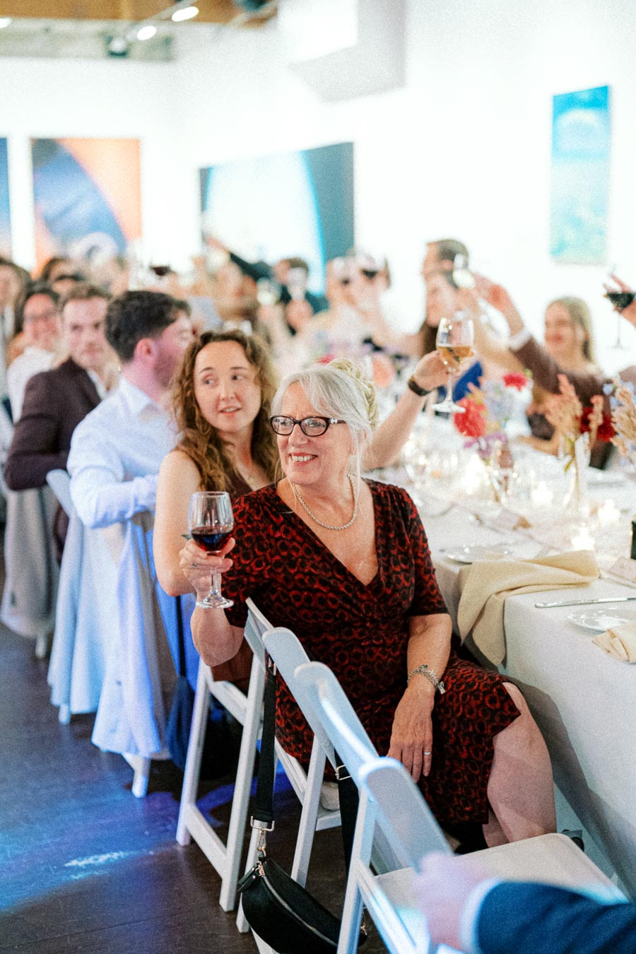 A joyful wedding reception scene with guests toasting, featuring a smiling older woman in a patterned dress holding a glass of wine at a beautifully set table, surrounded by other attendees in a bright, art-adorned venue.