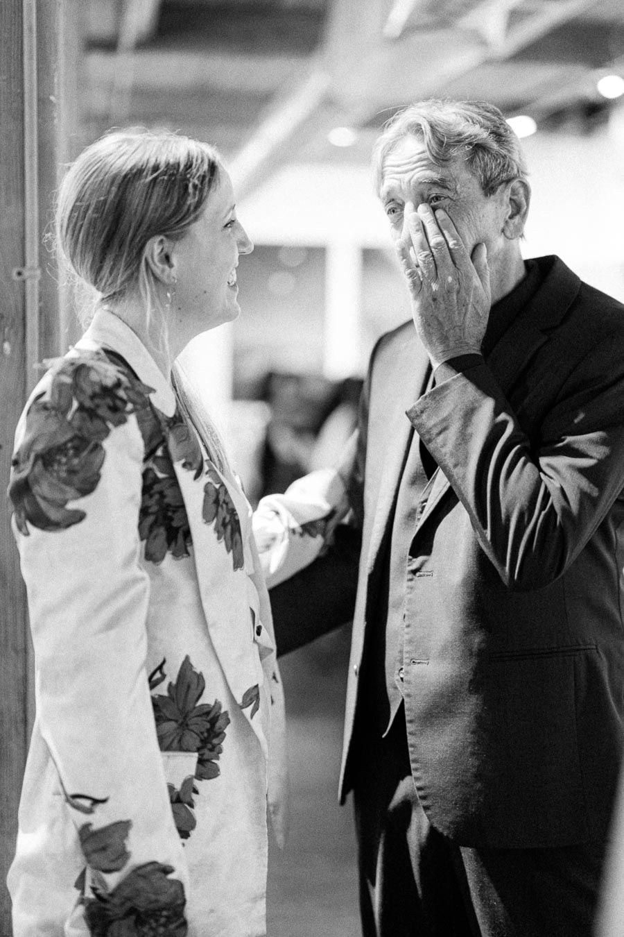 Black and white photo of a woman in a floral jacket and a man in a suit sharing a heartfelt moment indoors.