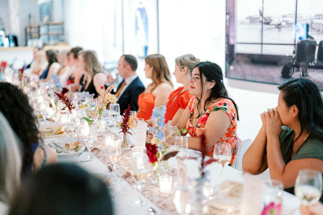 Guests seated at an elegantly decorated long dining table during a formal event, featuring vibrant floral centerpieces and softly lit candles, creating a warm and inviting atmosphere.