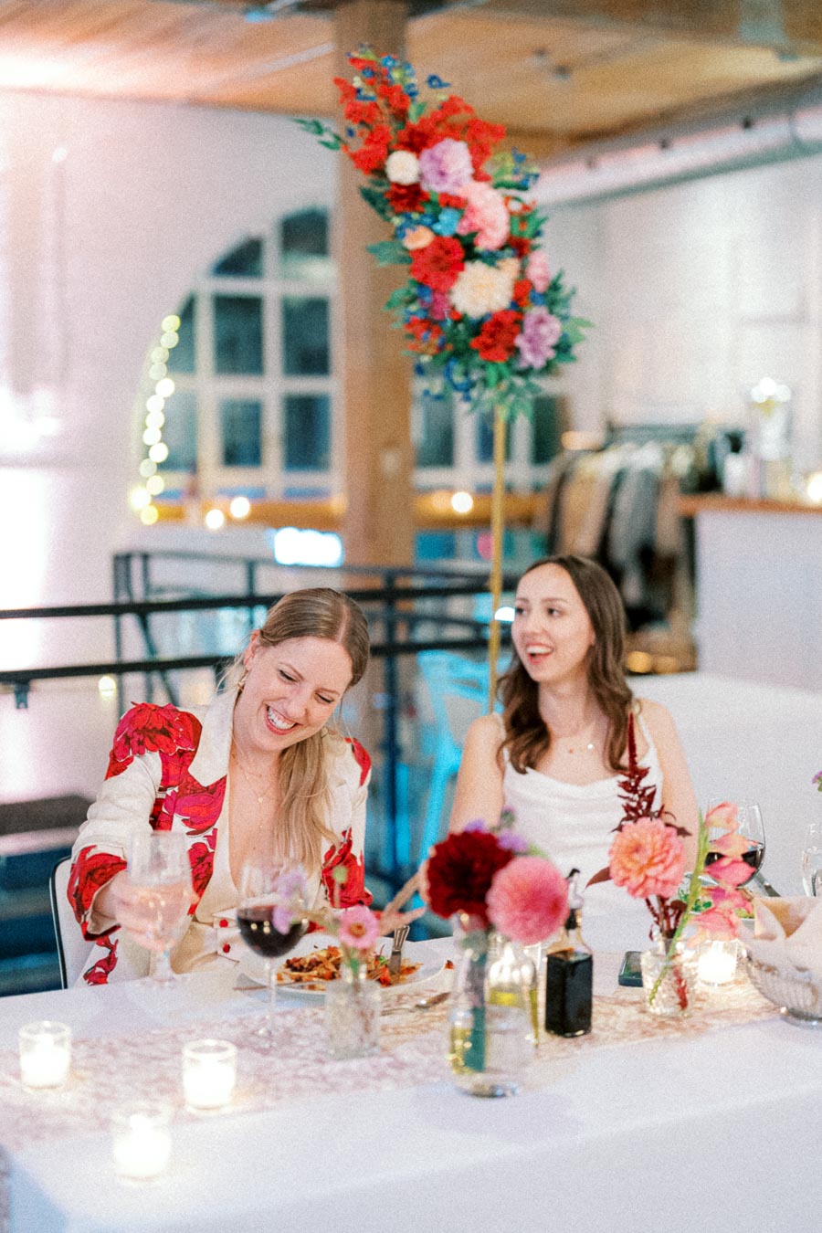 Festive indoor gathering with two women enjoying a meal at a decorated table adorned with colorful floral centerpieces and candlelight ambiance.
