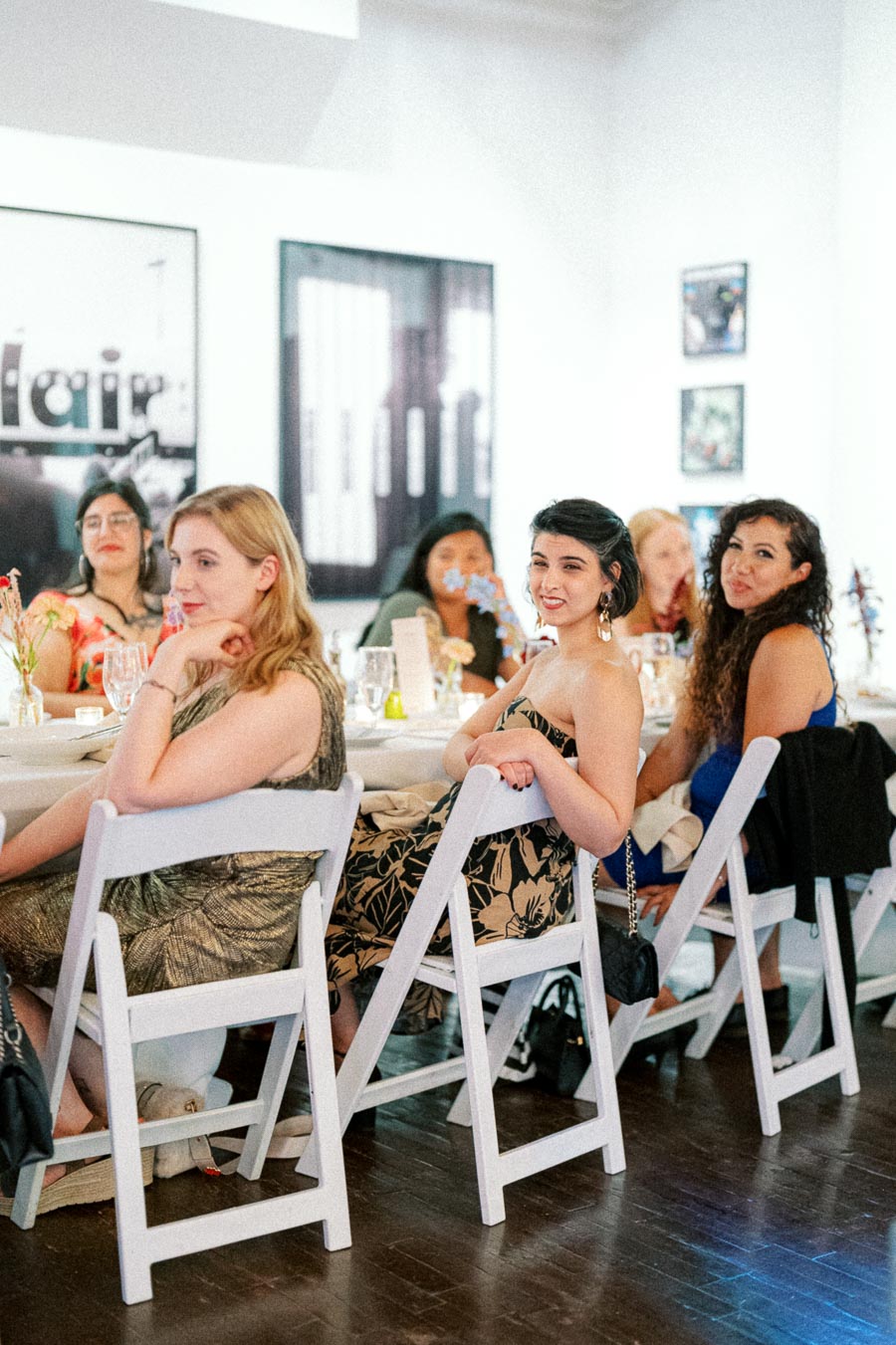 Group of elegantly dressed women sitting at a banquet table in a well-lit event space, engaged and smiling, with art pieces visible in the background.