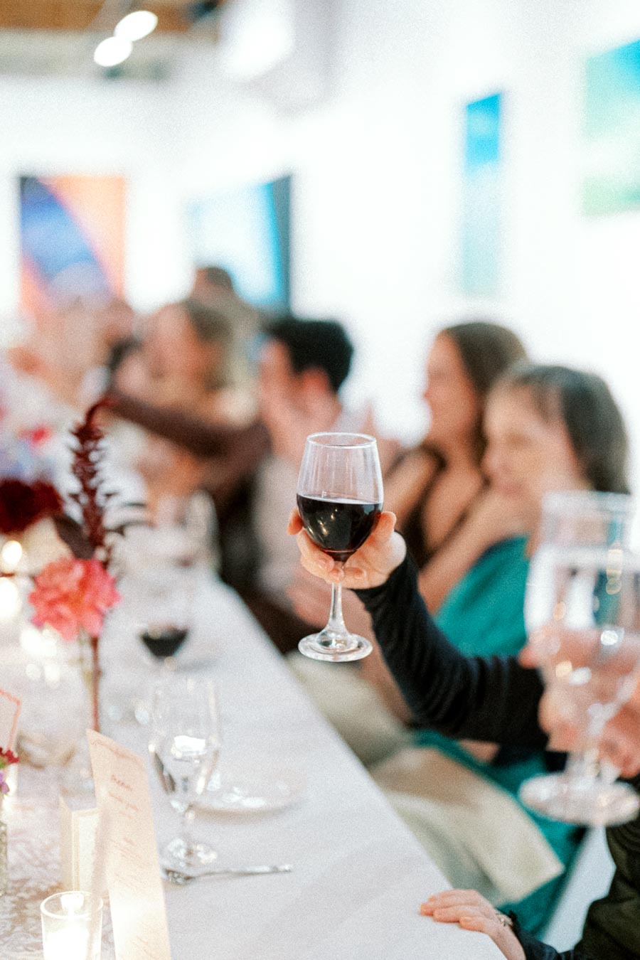 A close-up of a hand raising a glass of red wine at an elegantly set table, surrounded by blurred guests in a bright, modern dining setting, suggesting a festive or celebratory event.