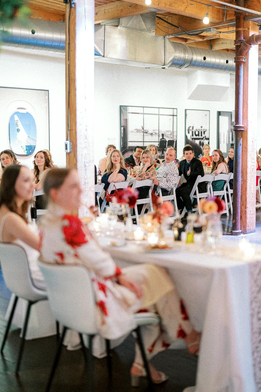 A group of people attentively listening during an indoor event, seated at tables with white chairs, surrounded by modern art and decorations in a spacious venue.