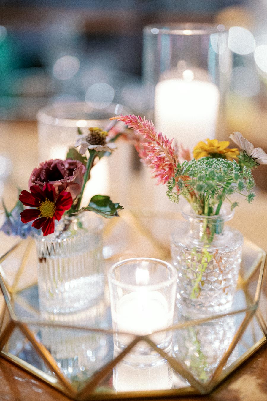 Elegant floral arrangement with vibrant flowers in glass vases on a mirrored tray, accompanied by a lit candle, creating a warm and cozy atmosphere.