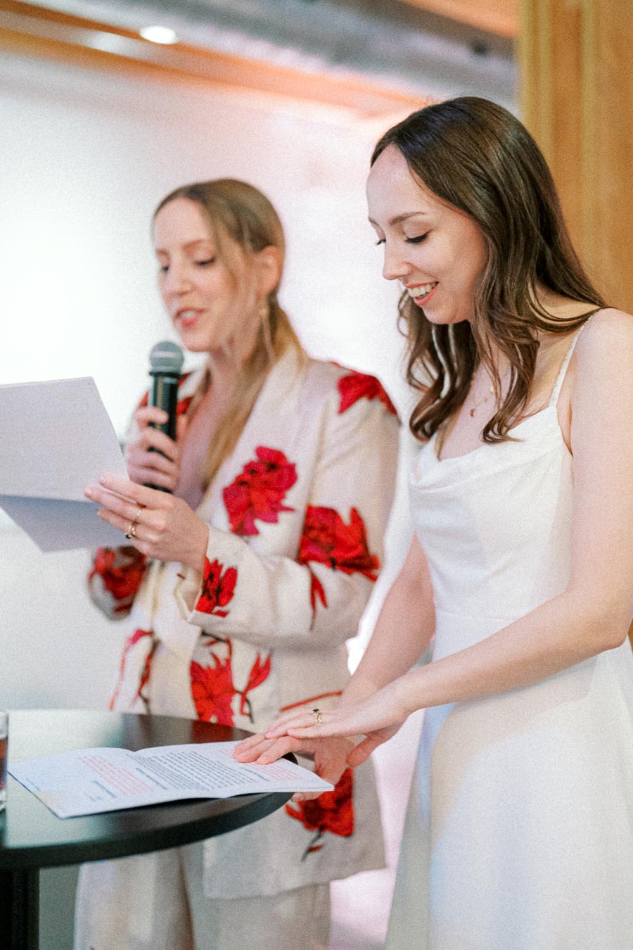 Two women at a formal event, one speaking into a microphone and both reading from papers, dressed in elegant outfits.
