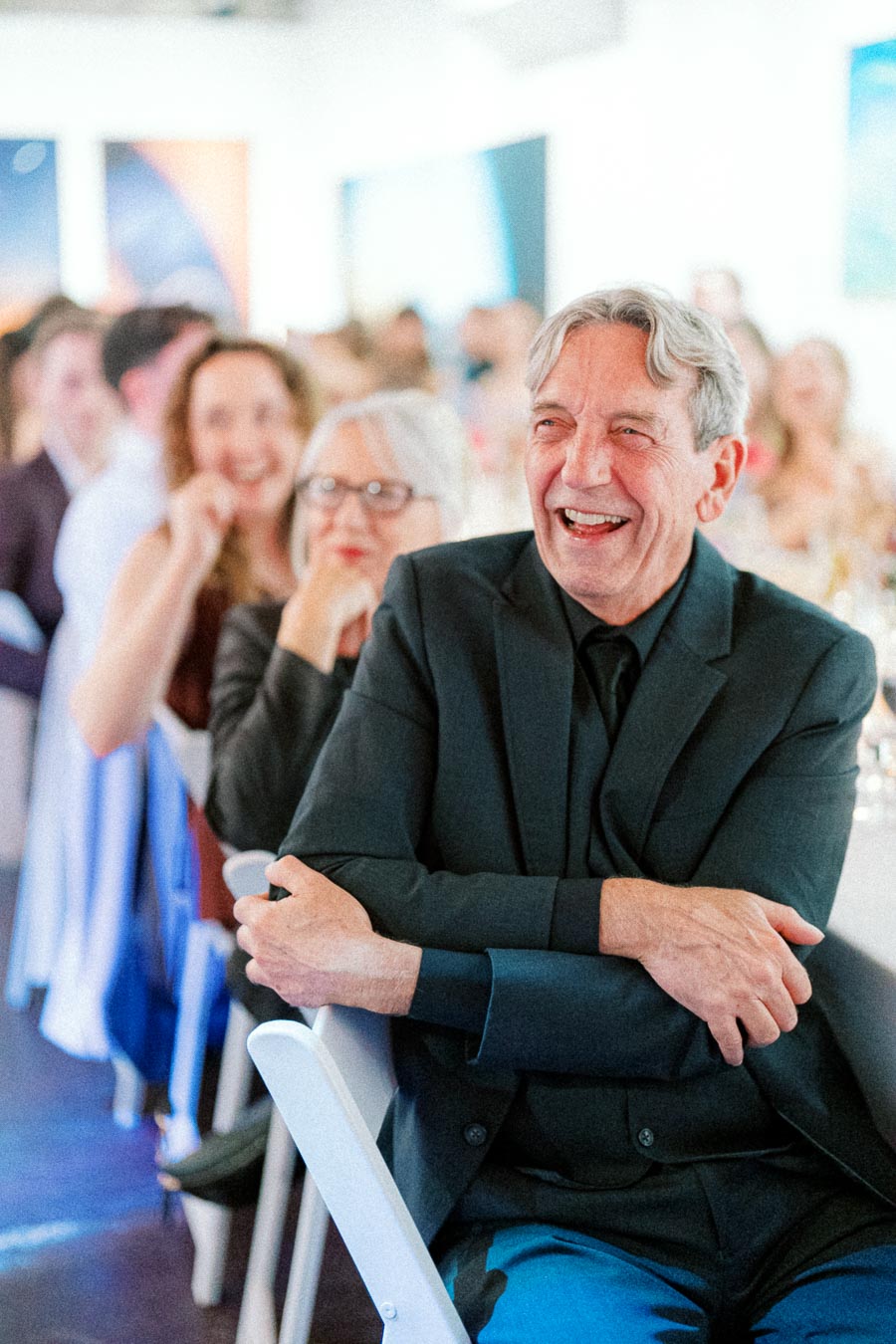 Elderly man in a black suit laughing joyfully at a formal event, with a blurred background showing smiling attendees seated at tables.