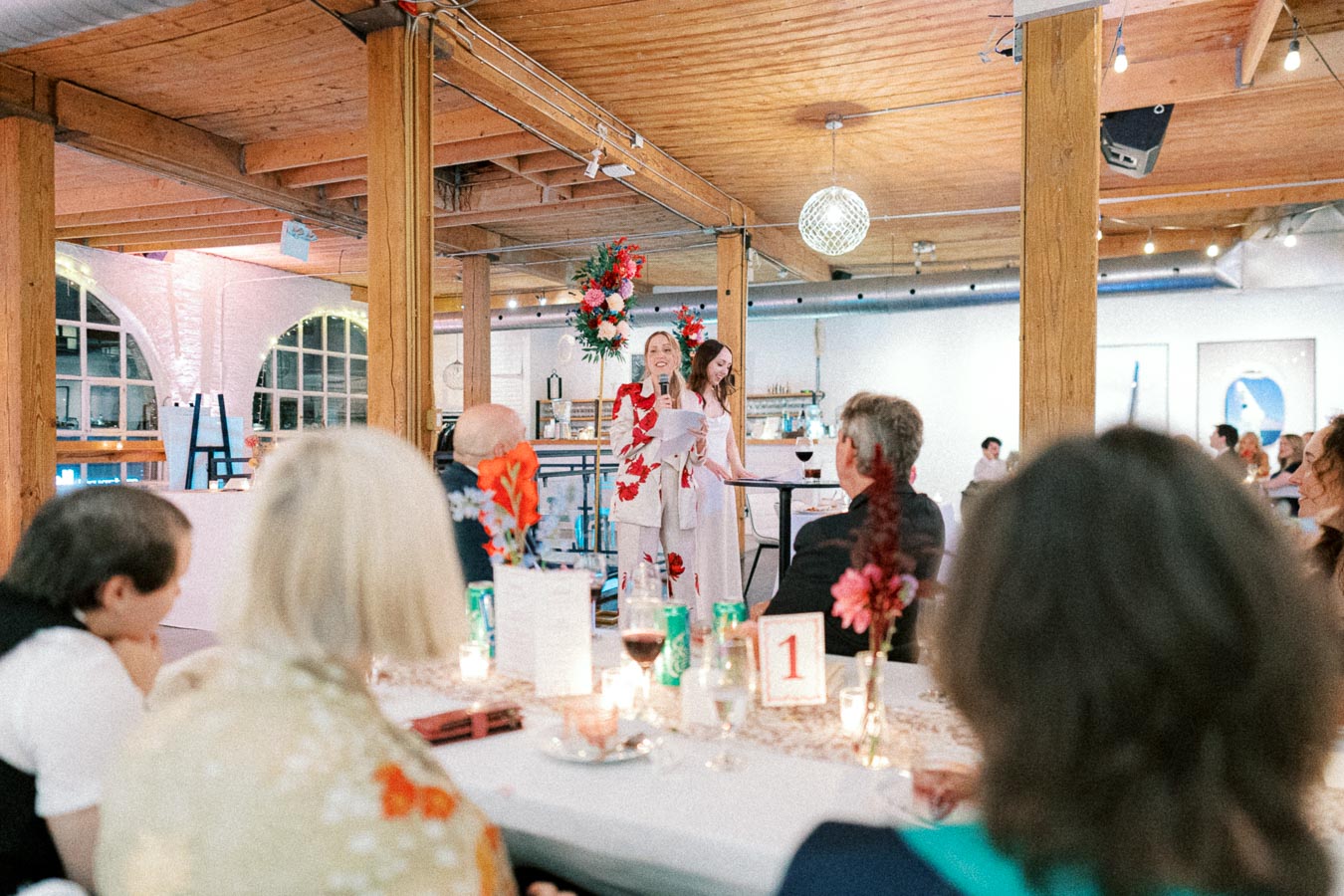 Guests sitting at a beautifully decorated table listen to a speech at a stylish indoor event with exposed wooden beams and ambient lighting.