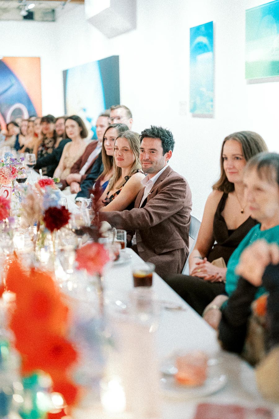 A group of people seated at a long table in an art gallery setting, engaged in a formal event with colorful flowers and artwork displayed on the walls.