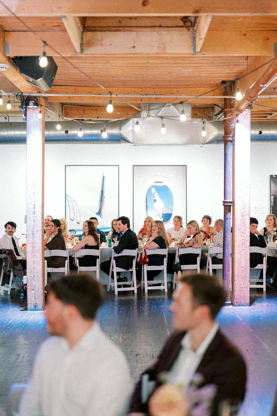 A group of people sitting at a long table in a modern event space with wooden beams and hanging lights, engaged in a formal gathering or celebration.