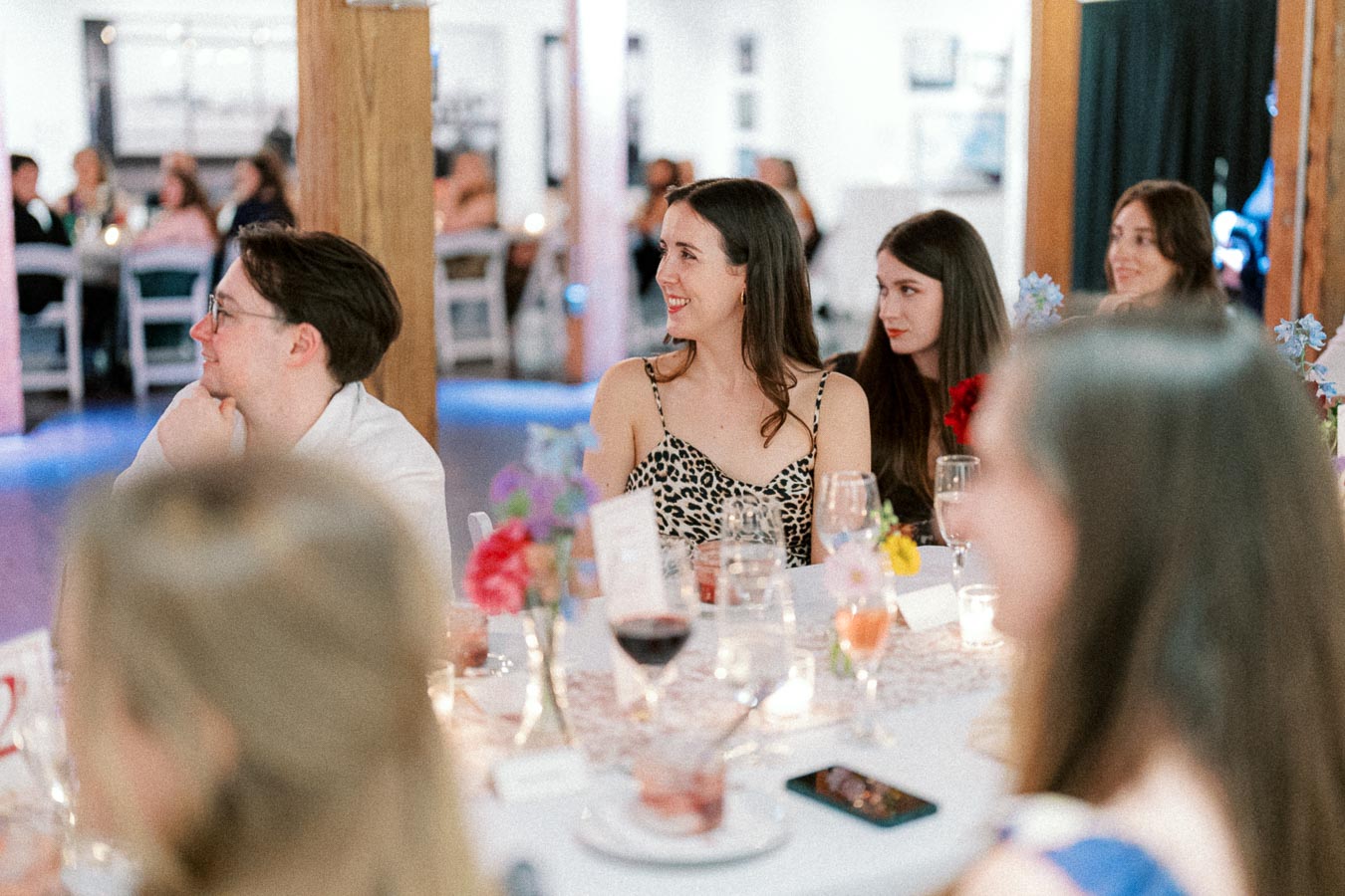 A group of people enjoying a social event seated at a decorated table with drinks and flowers, engaged in conversation and laughter.