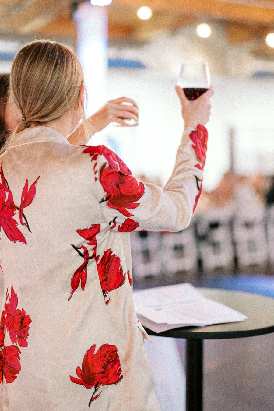 Woman in a floral jacket holds a glass of red wine during a social event, standing beside a table with documents in a well-lit setting.