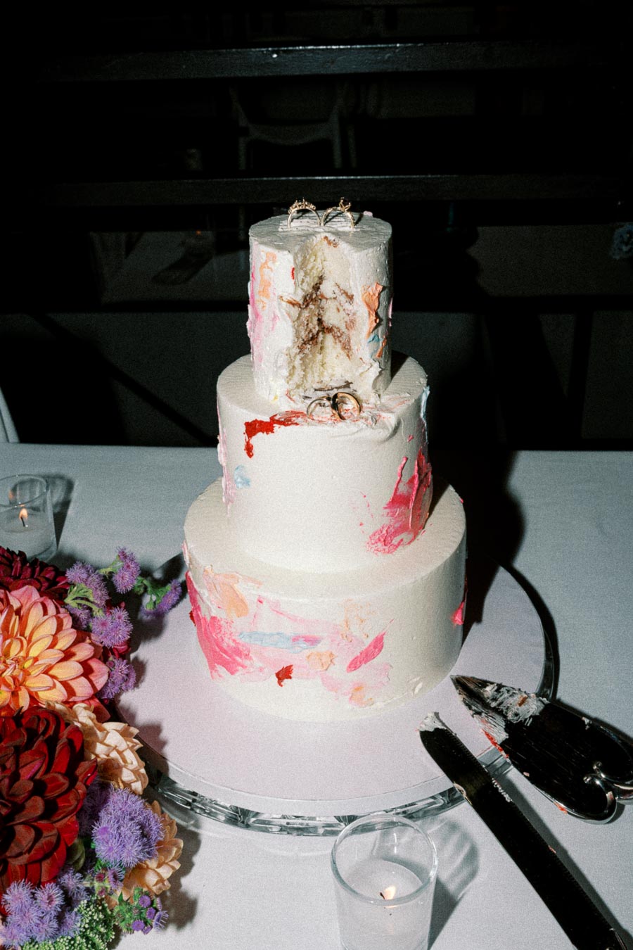 Three-tier wedding cake with colorful abstract icing design, adorned with two rings on top. Surrounded by vibrant flowers and candlelight, with cake-cutting utensils nearby on a white tablecloth.