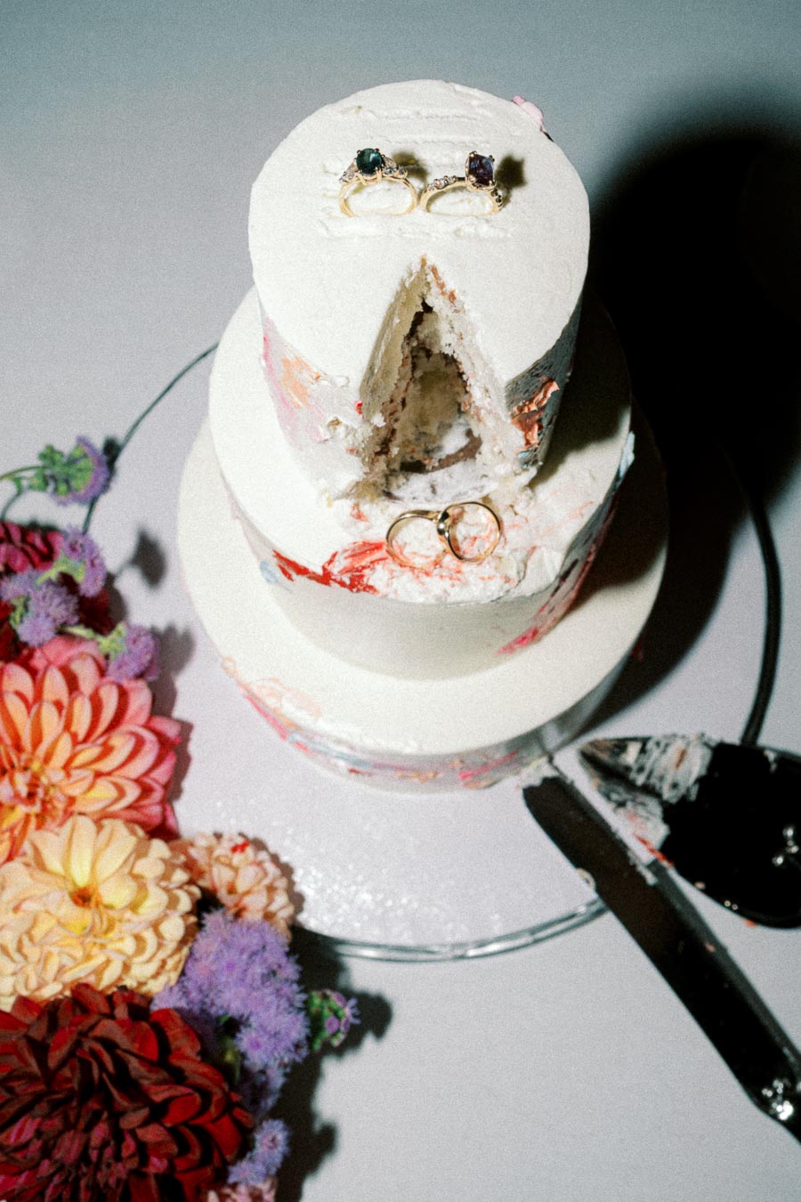 Two-tiered wedding cake with a slice removed, revealing rings placed on top, surrounded by vibrant flowers and a cake knife on the side.