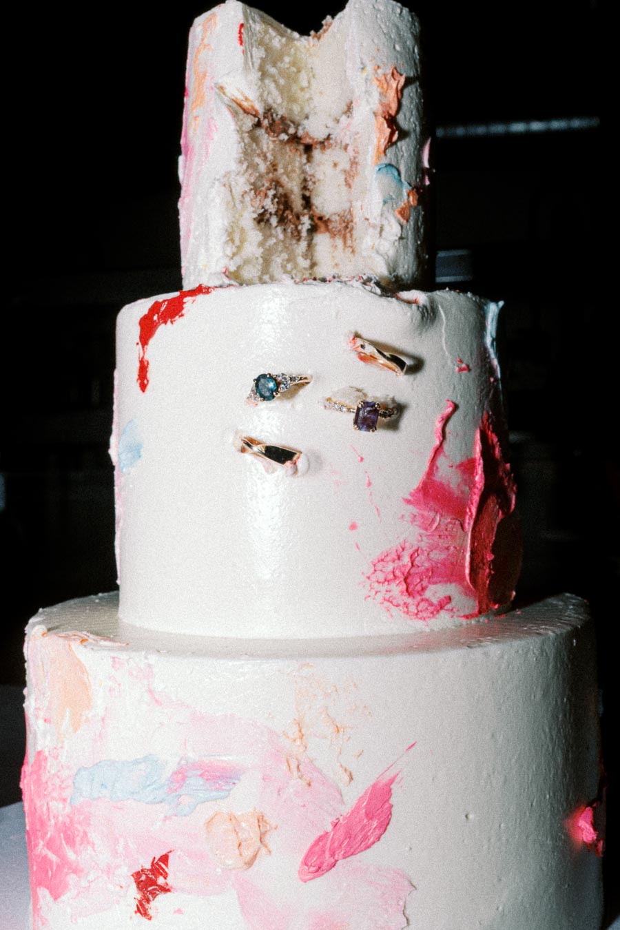 Three-tiered artistic wedding cake with colorful abstract paint splashes and embedded gemstone rings, featuring a partial bite on the top layer.
