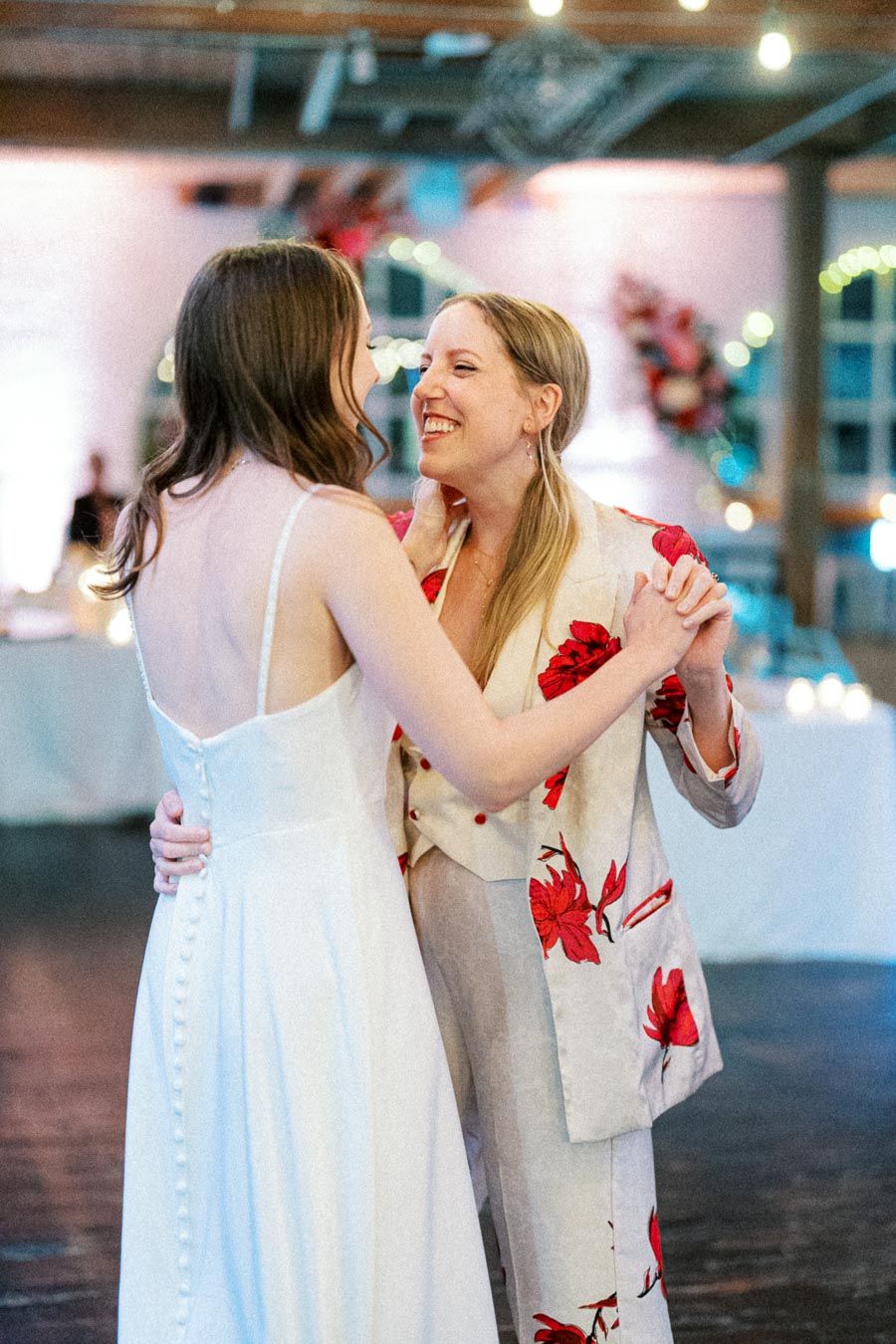 Two people embracing at a wedding reception, one wearing a white dress and the other in a floral-patterned suit, smiling joyfully in a beautifully decorated venue with soft lighting.
