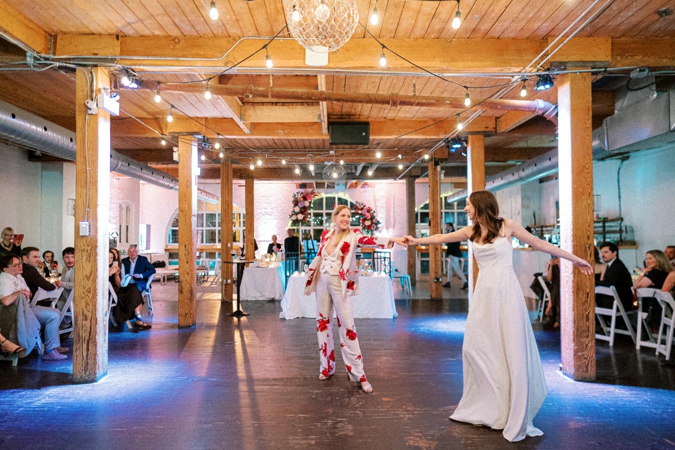 A joyful couple dancing at their wedding reception in a warmly lit rustic venue, with wooden beams and elegantly dressed guests seated at tables in the background.