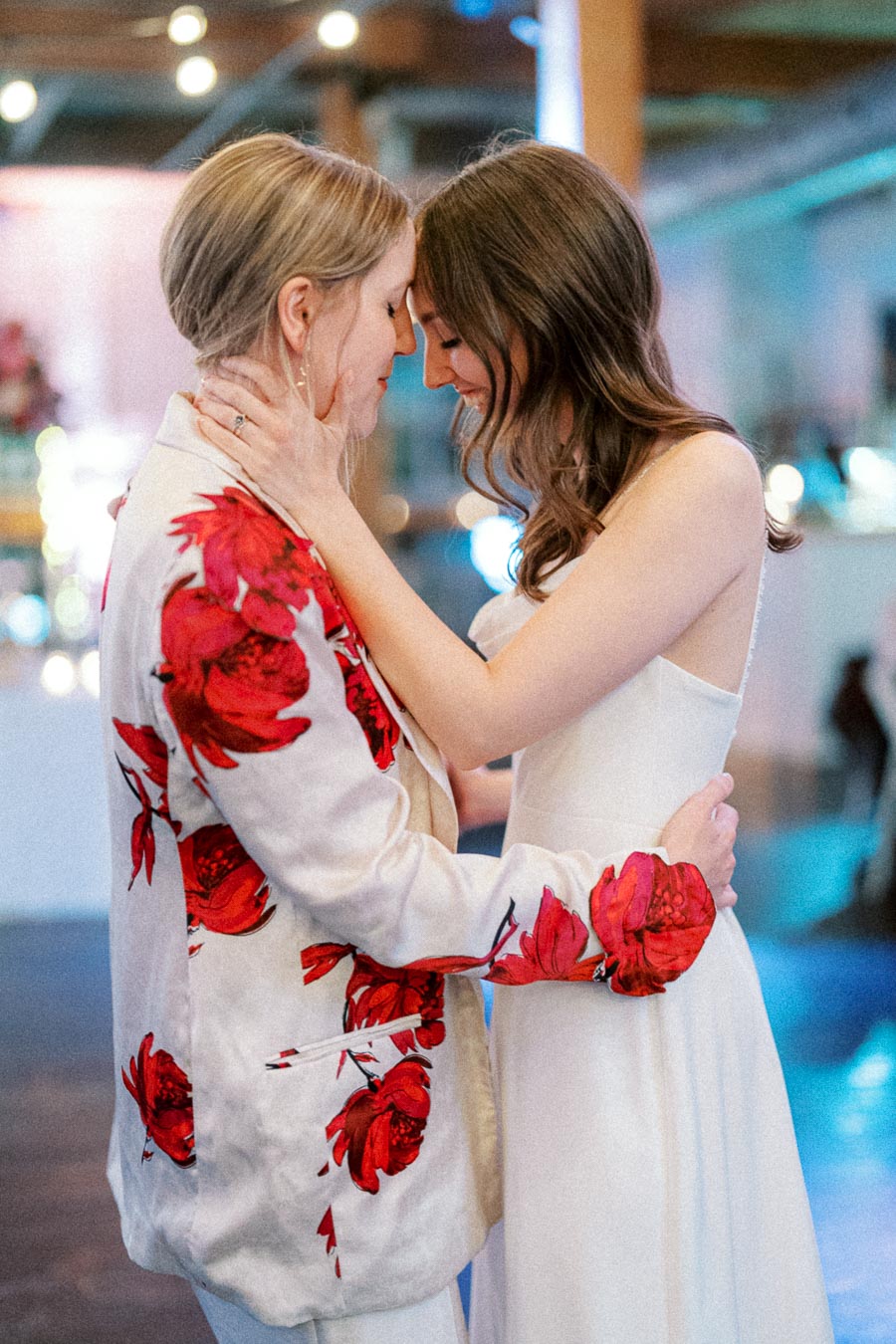 A romantic, intimate wedding moment between two brides, one in a white floral-patterned suit and the other in a strapless white gown, embracing with foreheads touching in a softly lit venue.