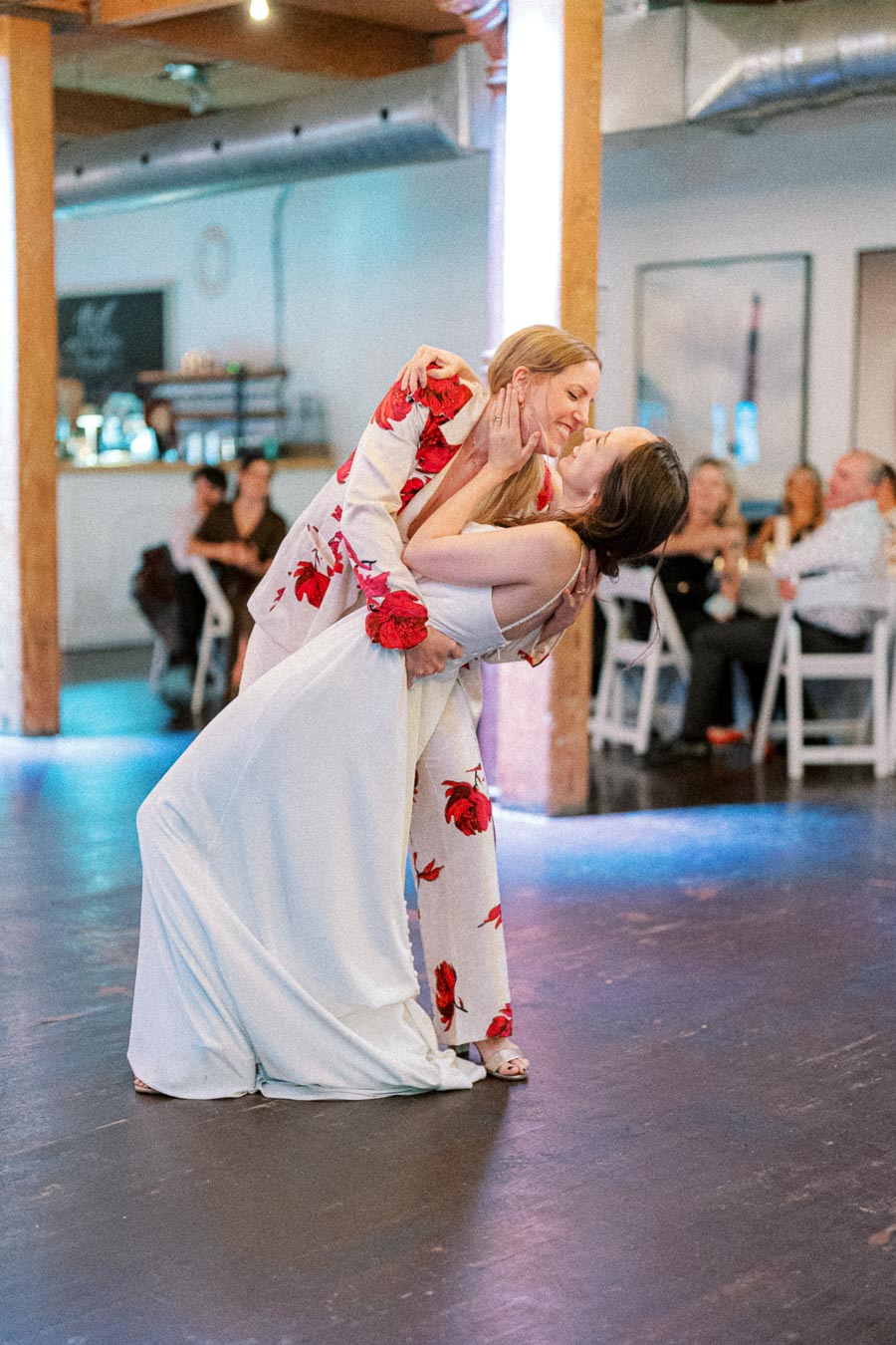 Two women sharing a romantic dance at a wedding reception, one in a white gown and the other in a floral suit, surrounded by seated guests in a warmly lit venue.