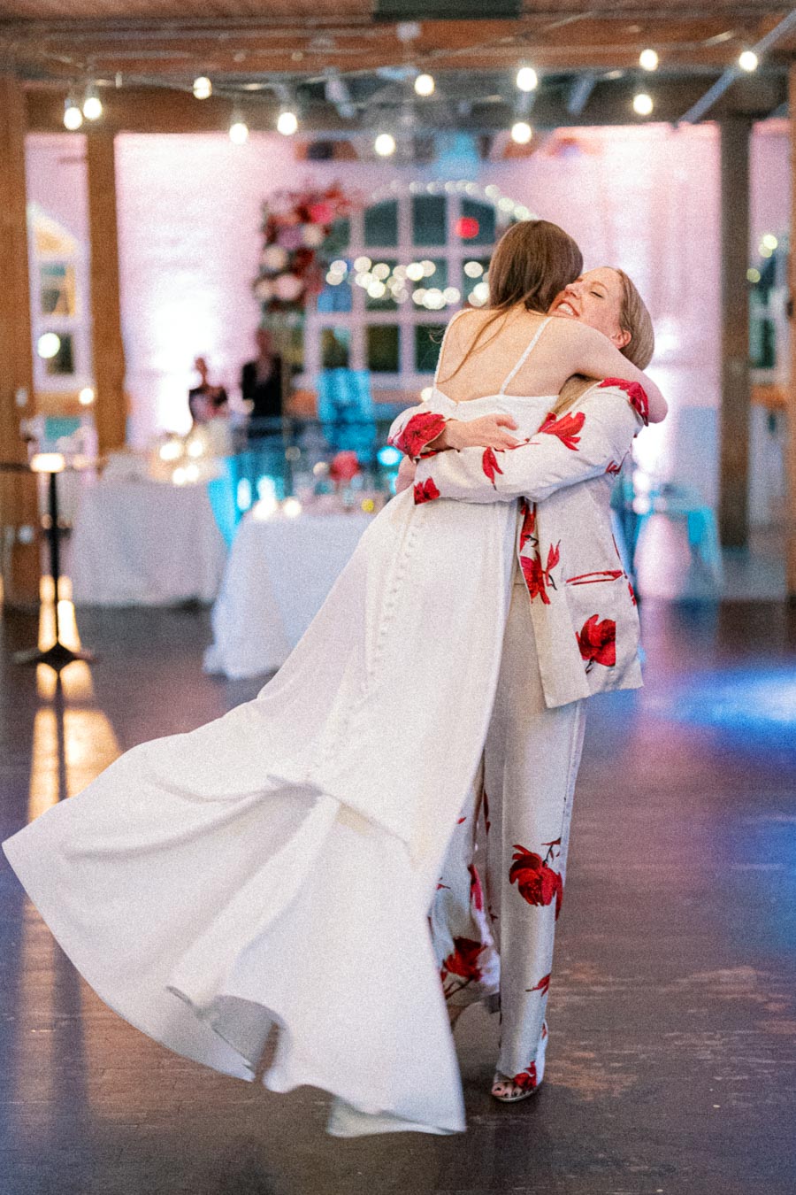 Two women embrace joyfully in a warmly lit wedding reception venue, one in a flowing white dress and the other in a floral-patterned outfit, surrounded by soft lighting and festive decorations.