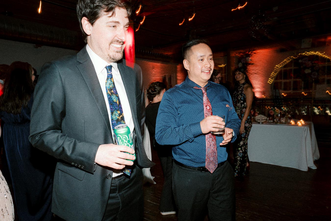 People socializing at a lively gathering, dressed in formal attire, with festive lighting and decor in the background.
