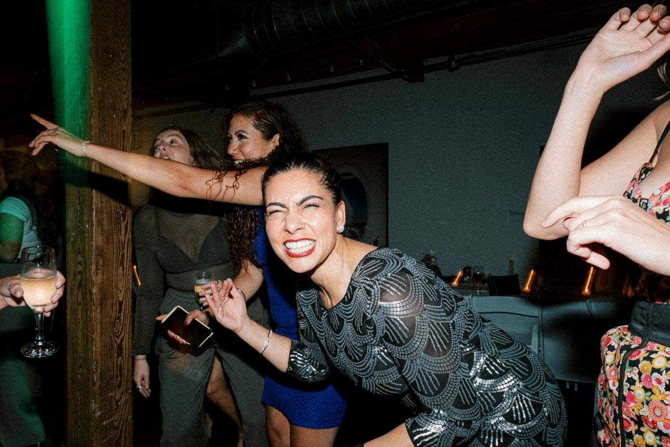 A group of women enjoying a lively party, smiling and dancing enthusiastically in a dimly lit venue, one wearing a patterned dress and holding a drink.