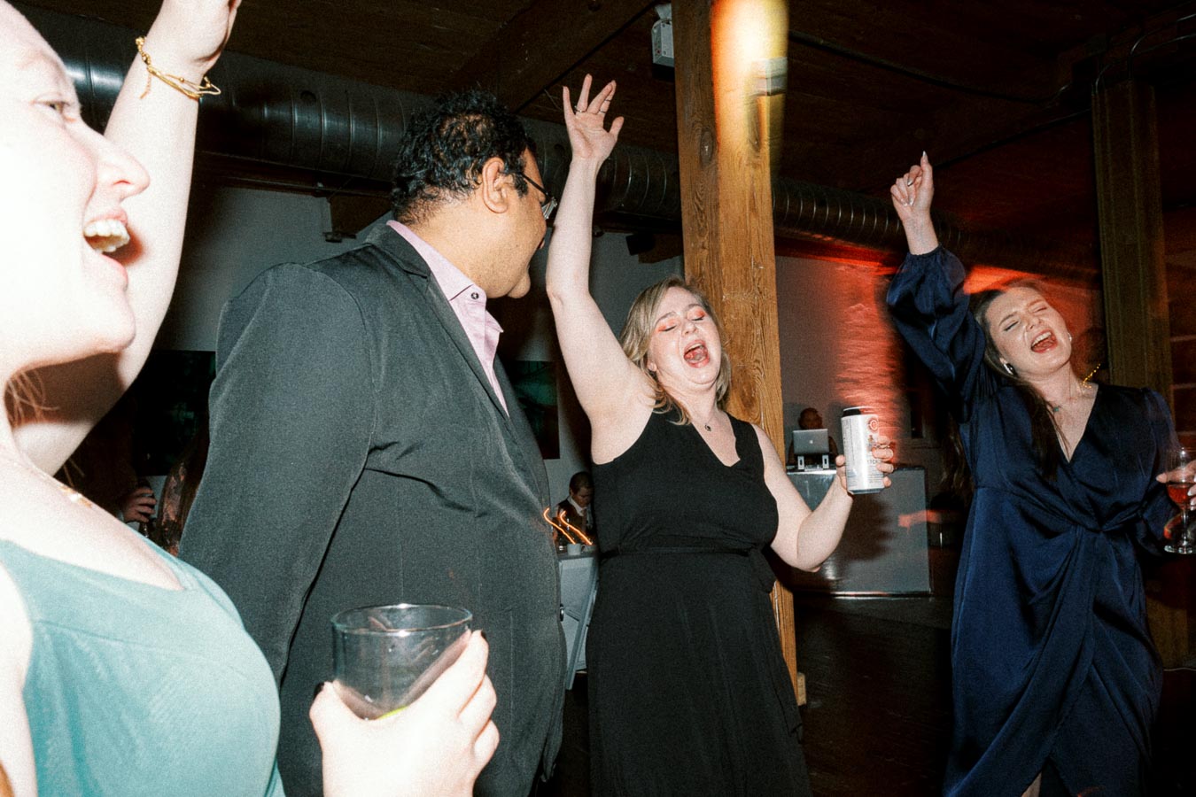 A group of people enjoying themselves, dancing and holding drinks, at a lively party in a dimly lit venue with exposed wooden beams.