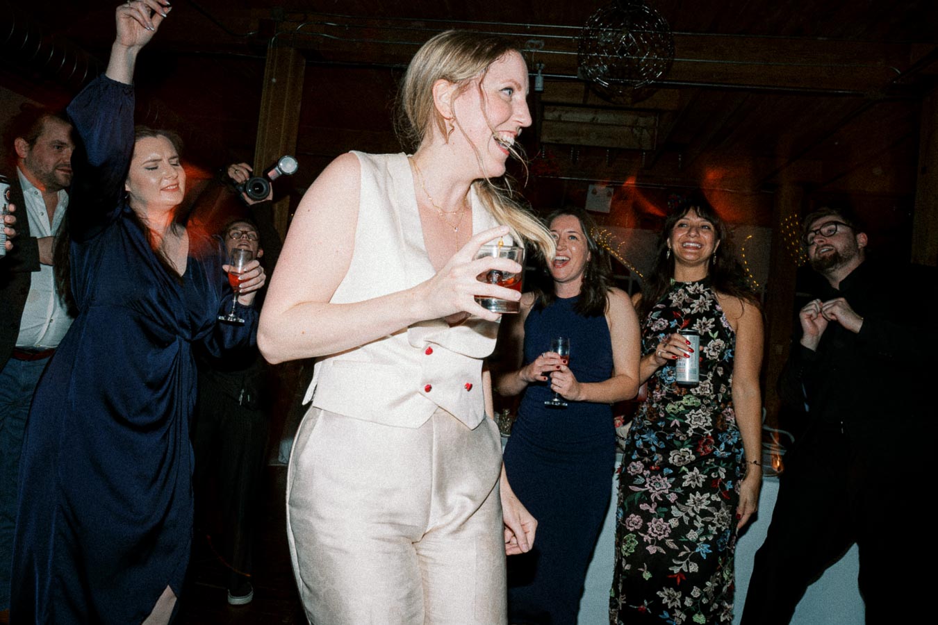 A group of people joyfully dancing at a party, with a woman in the foreground holding a drink and others smiling and holding beverages, capturing a lively celebration atmosphere.