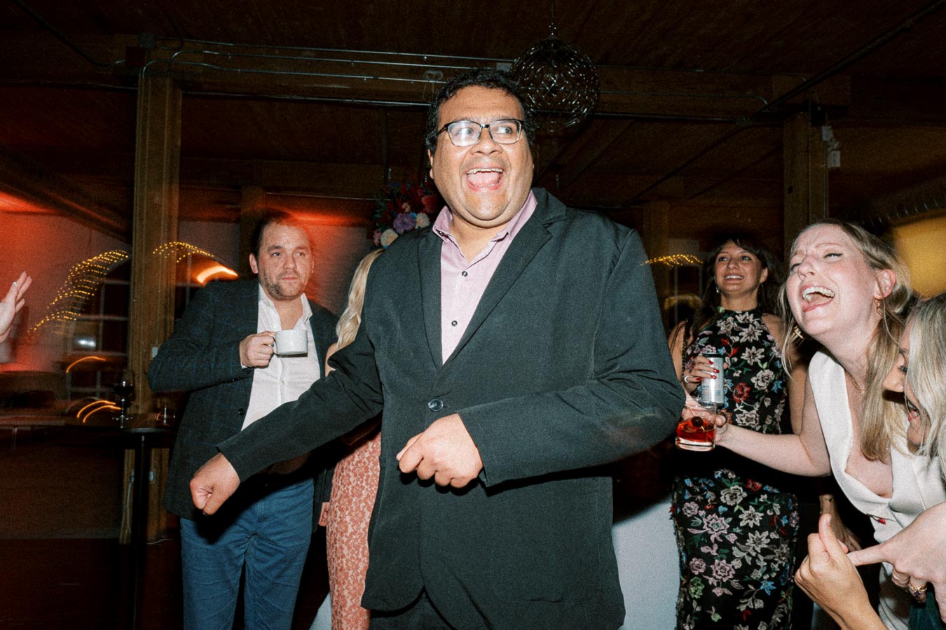 Group of people joyfully celebrating at an indoor party, with one man in a suit leading the laughter while others with drinks join in the fun, under the warm glow of ambient lights.
