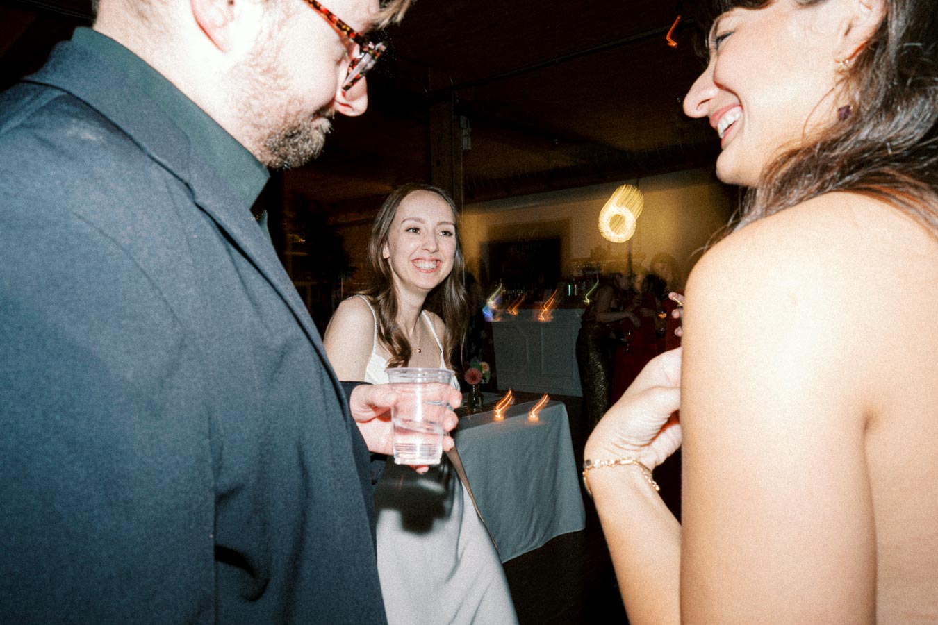 A group of people enjoying a lively party with drinks in hand, captured in a warmly lit indoor setting. The atmosphere is celebratory with guests smiling and engaging in conversation.
