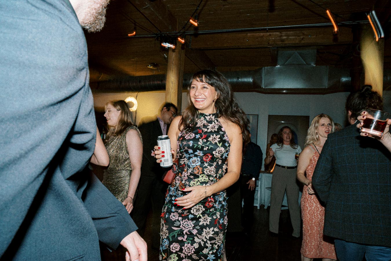 A woman in a colorful floral dress smiles and holds a drink can at a lively indoor party, surrounded by people socializing and dancing under warm ambient lighting.