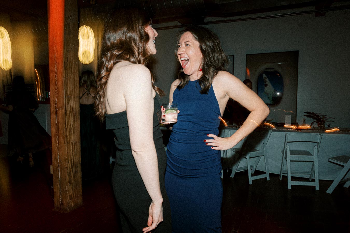 Two women dressed in elegant evening wear, laughing and enjoying drinks at an indoor party with a festive atmosphere.