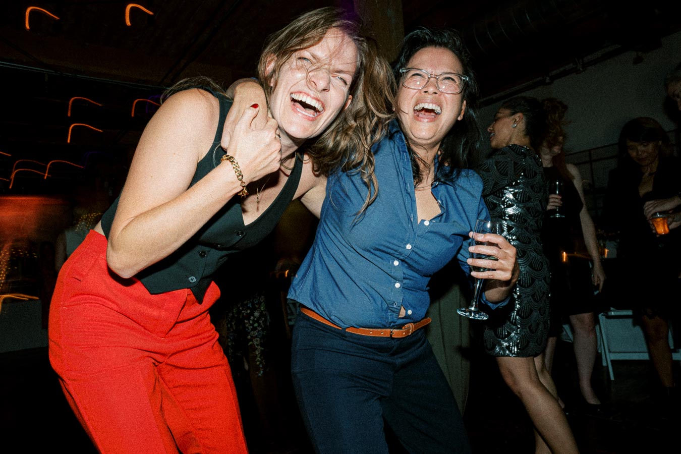 Two friends laughing and enjoying a night out at a lively party, with one holding a glass in their hand, surrounded by a joyful crowd.