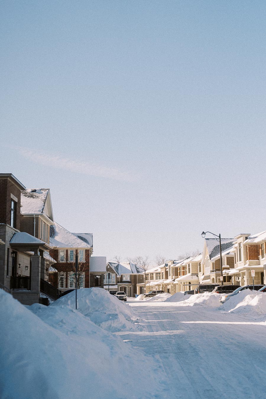 Snow-covered suburban street lined with modern houses, clear blue sky, winter morning scene.