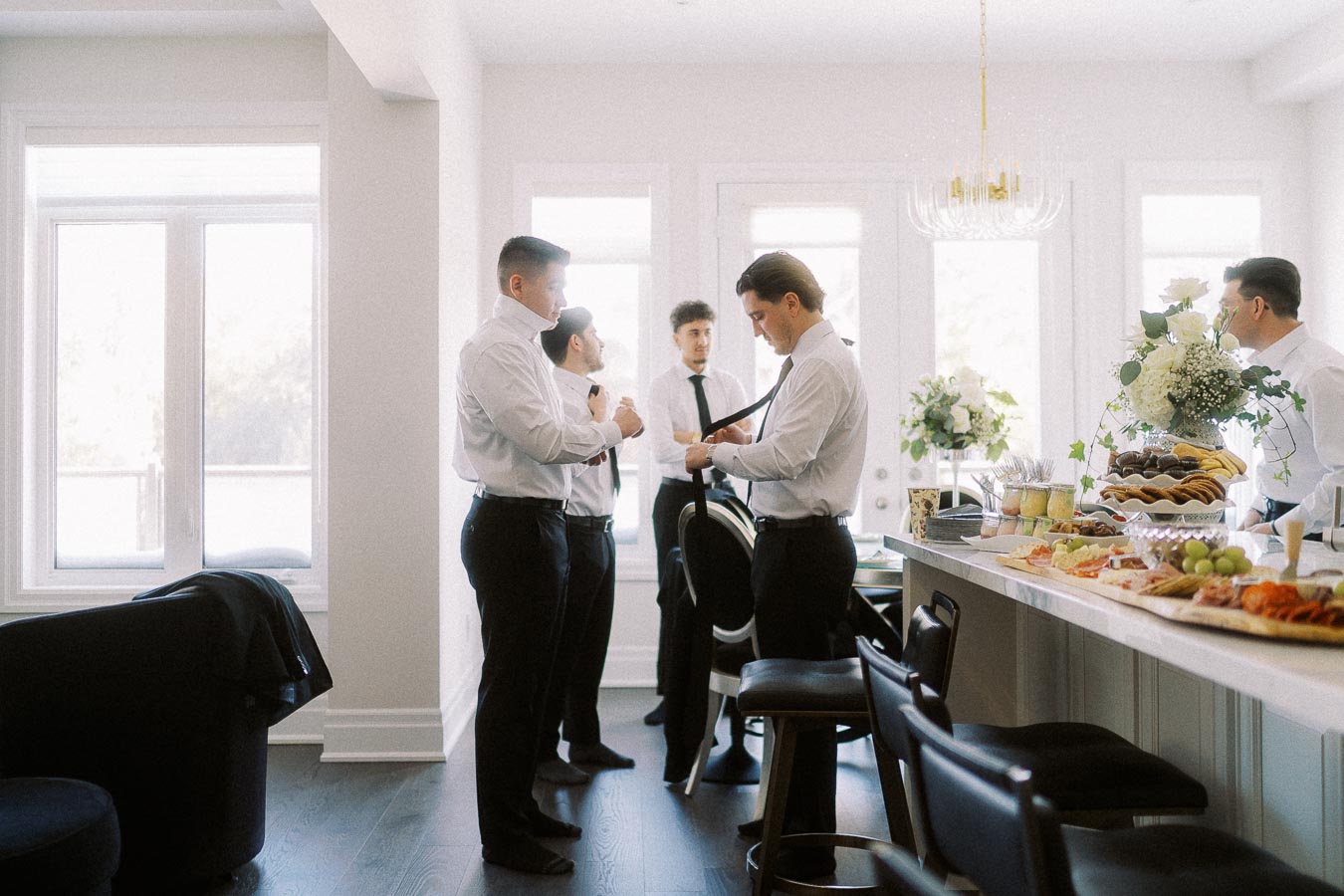 Groomsmen preparing for a wedding in a bright room, adjusting ties and jackets near a kitchen counter adorned with flowers and a variety of appetizers.