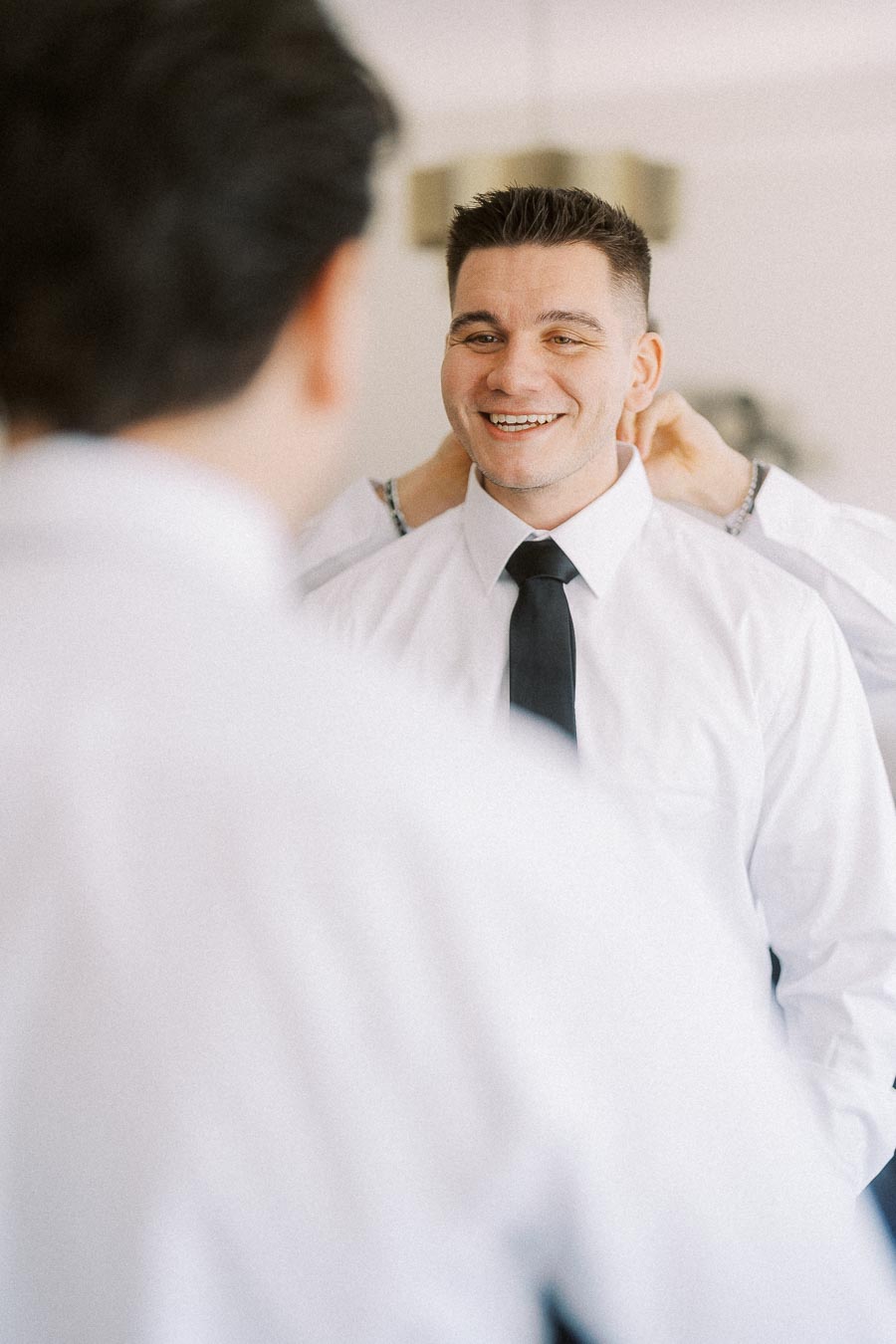 Young man smiling while adjusting his black tie in front of a mirror.