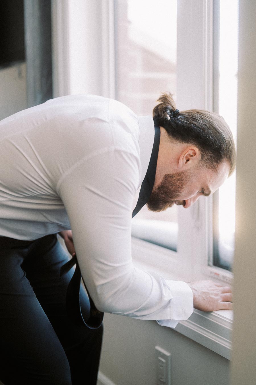 Man in a white dress shirt and dark pants leaning over a window sill indoors, adjusting something.