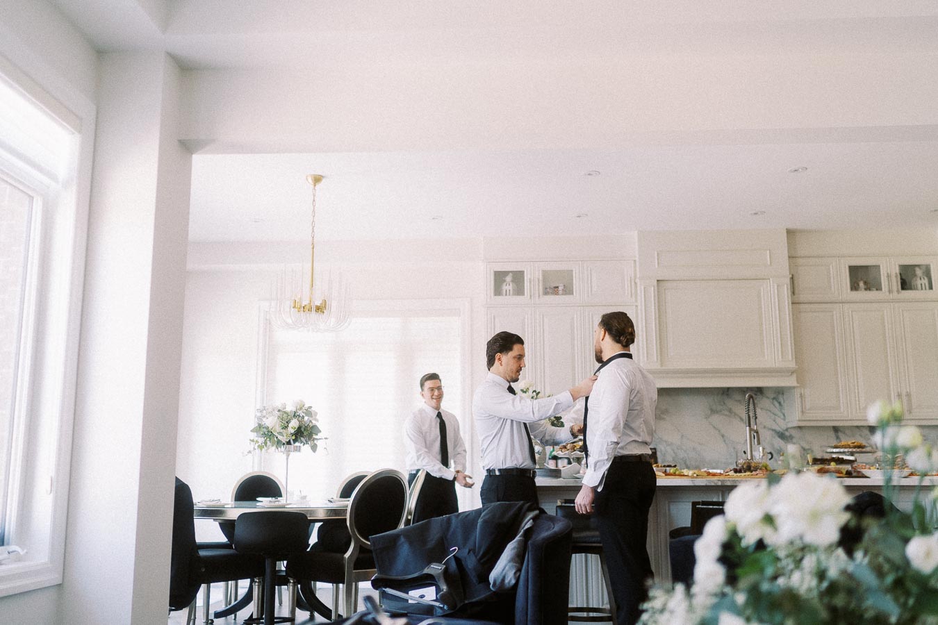 Groomsmen preparing for a wedding in a bright kitchen, with one helping the other adjust a tie, surrounded by elegant decor and floral arrangements.