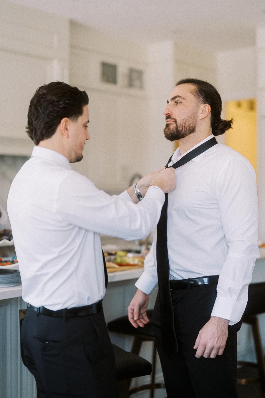Two men in formal white shirts adjusting a black tie, preparing for an event in a modern kitchen setting.