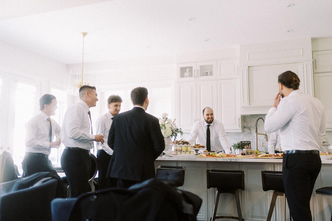 Group of men in formal attire socializing in a bright kitchen, enjoying food and drinks set on a marble countertop.