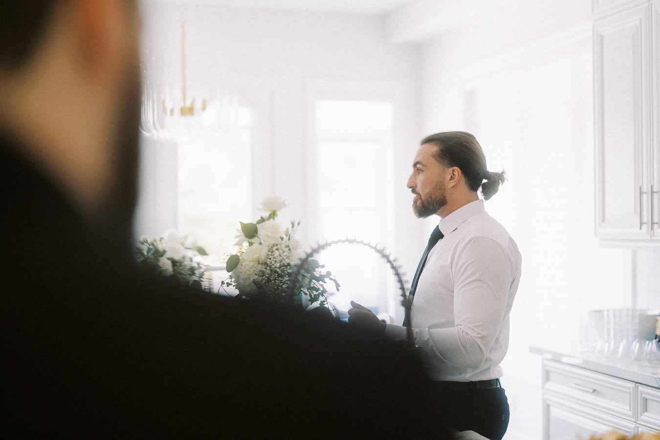 Man in a white shirt and black tie standing in a kitchen with flower arrangements and modern decor.